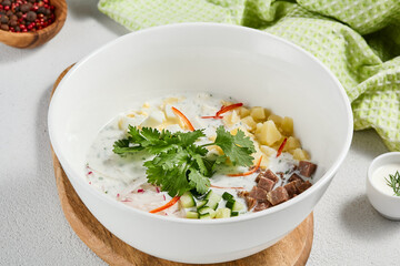 Traditional russian soups - okroshka with fresh vegetables, meat and kefir. Cold summer soup on white table. Aesthetic composition with traditional russian food and spices, textile