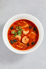 Fish tomato soup with shrimps and black olives. Seafood soup with prawns in white ceramic bowl on concrete background. Fish dish with seafood. Tomato soup in minimal composition.