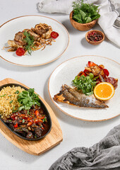 Set of main course: meat with bulgur, fish and vegetables, meat medallions on mashed potatoes. Composition with hot dishes on ceramic plate and wooden board. Food menu. Set with meat and fish dishes.