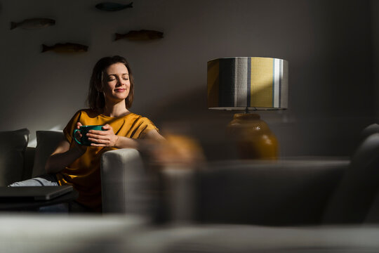 Smiling woman with eyes closed holding coffee mug sitting in living room