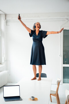 Happy businesswoman wearing headphones enjoying music standing on desk in office