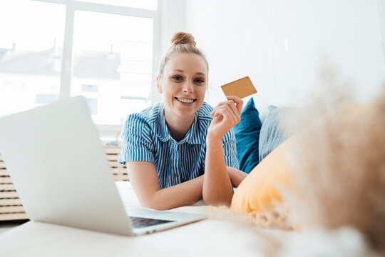Smiling freelancer showing credit card by laptop lying on bed at home