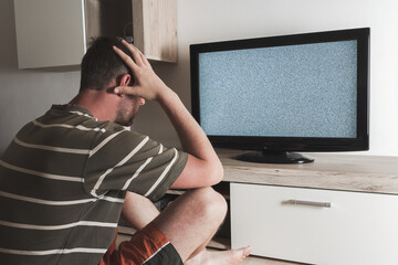 Young caucasian man sitting with head in hands looking to grey graining television. Depression,...