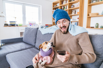 Shocked man with wooly hat sitting on couch holding dog checking smartphone
