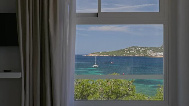View throught the window of a room in a hotel with sea, trees, mountains, yachts and a boat floating on turquoise water. Ibiza, Spain.