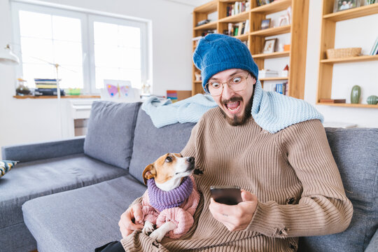 Shocked man with wooly hat sitting on couch holding dog checking smartphone