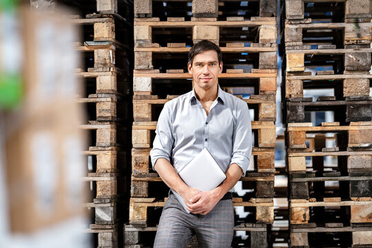 Confident businessman with laptop in front of stack of pallets at warehouse
