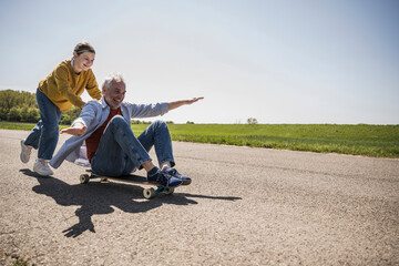 Playful granddaughter pushing happy grandfather sitting on skateboard