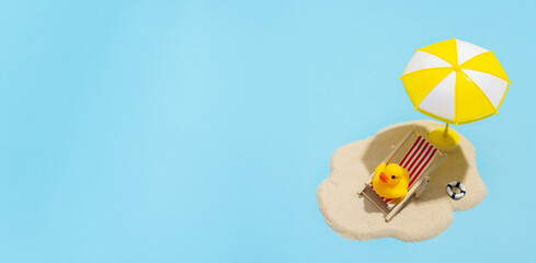 Summer concept. Sun umbrella and duck in sun lounger on sea sand on blue background. Sun protection...