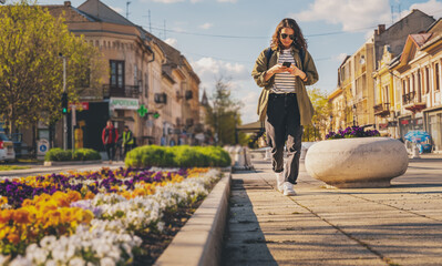 Fototapeta na wymiar Stylish young woman in sunglasses walks looking into a smartphone on the street in a summer European city
