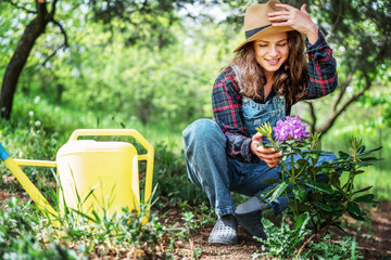 Beautiful young woman gardener in a hat with a watering can looking at a hydrangea bush in her...