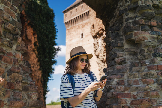 Young cheerful woman traveler in a hat and with a smartphone in her hands against the backdrop of the ruins of a medieval castle