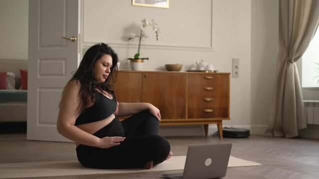 Young overweight woman doing yoga in front of laptop and sitting on floor at home spbd. 4k Beautiful caucasian female does asana or exercise and looks at computer, trains calmly and sits on mat in