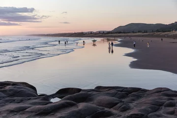 Foto op Canvas Zonsondergang Strand Muriwai beach in the pinkish colours of the setting sun. Unrecognizable silhouettes of several people spending the evening on the beach.   © Anna J. Photography