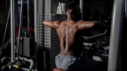 Fototapeta na wymiar Shirtless african american man doing back exercises on a machine in the gym.