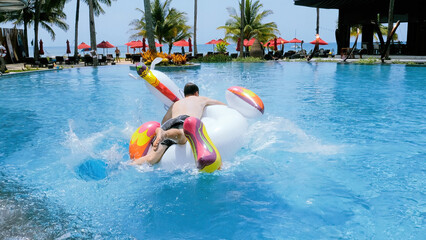Happy funny man successfully jump onto floating inflatable unicorn in swimming pool in tropical...