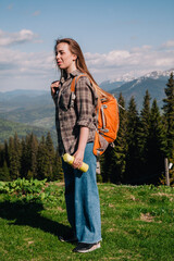 Naklejka premium A young, girl in a plaid shirt and jeans with loose hair with an orange backpack drinks tea from a bright thermal cup against the backdrop of the mountains in the Carpathians. view from the back.