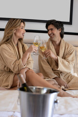 Handsome couple in bathrobe sitting on the hotel bed toasting and drinking champagne