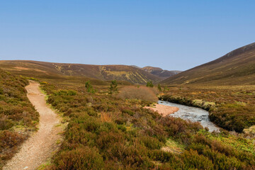 Fototapeta na wymiar A scenic view of a Scottish moutain river (Nethy) with moorland and path in the foreground and mountain valley and summit in the background under a beautiful blue sky