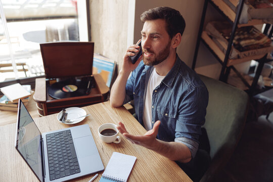 Caucasian handsome man talking on cellphone in cafe while sitting with coffee and laptop. Phone call