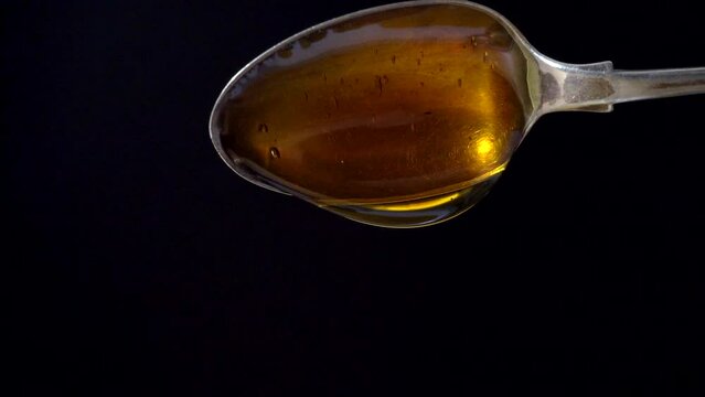 Closeup, isolated on black, of thick, sweet, smooth, translucent, amber syrup / light treacle, slowly pouring from a large spoon.