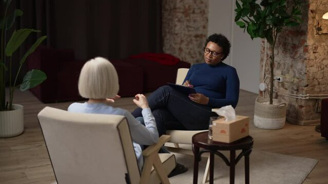 Female psychologist is listening to patient and writing while sitting in chair in office spbas. 4k Young American African woman listens to story and writes on clipboard, elderly lady speaks and sits