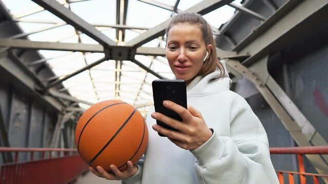 Young woman basketball player with headphones holding ball using smartphone after training. Urban background.