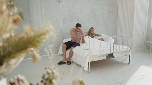 A Happy Young Family Lies Down In A White Bed With A Good Mood. Blonde Mom Covers Her Son Sitting In Bed.