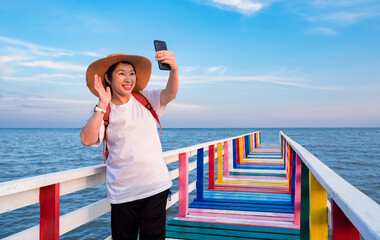 Happy Asian female tourist waving hand while video call with  smartphone on colorful wooden bridge at sea viewpoint against blue sky background