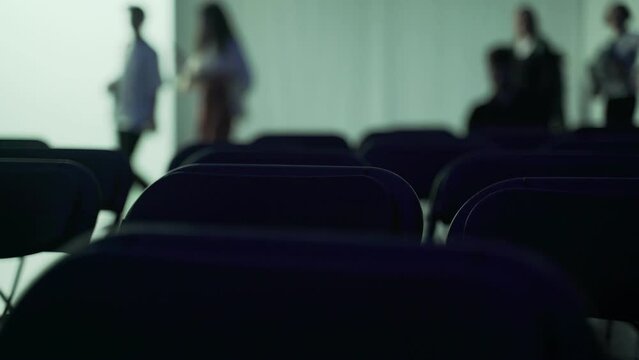 Public event. Concert, lecture, talk. Close-up of the backs of chairs arranged in even rows. Silhouettes of chairs on a background of colored light. Blurry silhouettes of people in the distance.
