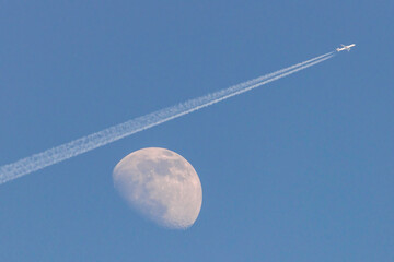 Biały księżyc w dzień na błękitnym bezchmurnym niebie. Nad księżycem przelatuje samolot...