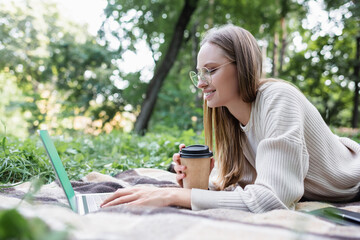 happy freelancer in glasses holding paper cup while lying on blanket and using laptop in park.
