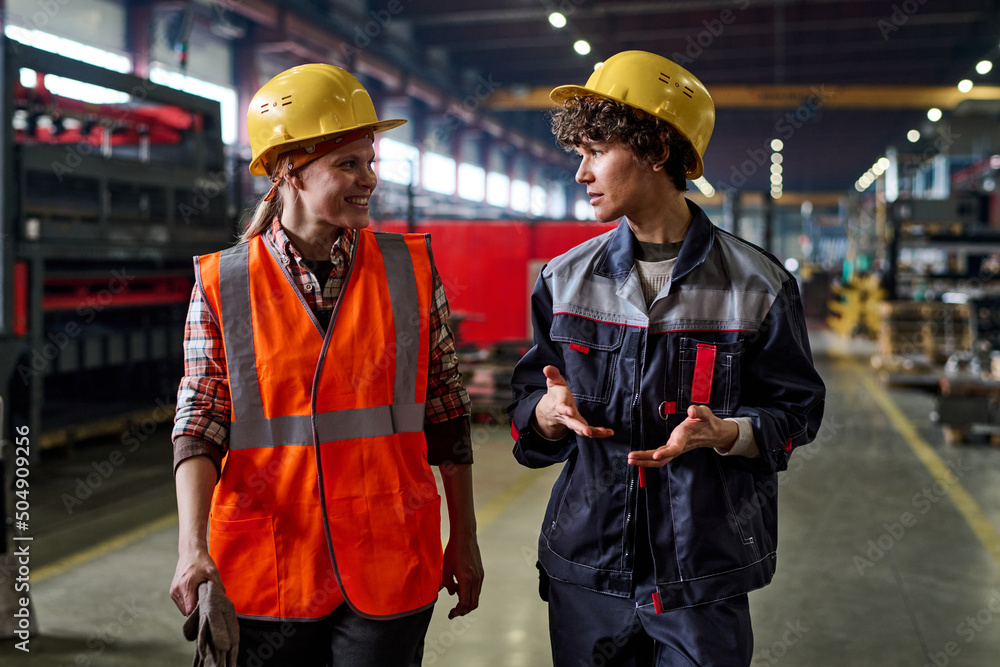 Aufkleber two young female workers of modern factory discussing working points while moving down large worksho