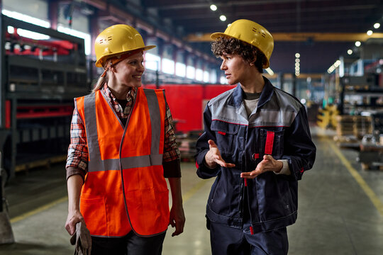 Two young female workers of modern factory discussing working points while moving down large workshop or distribution warehouse