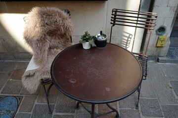 Runder Bistrotisch und alter Bistrostuhl mit Lammfell auf dem Bürgersteig vor einem Café und...