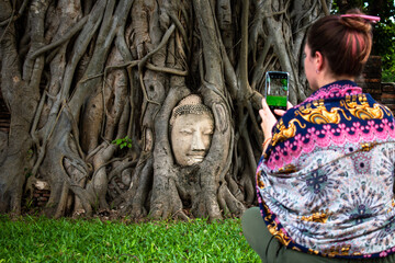 Mujer turista fotografiando cabeza de buda en ruinas, en raíces de un árbol antiguo