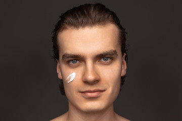 Facial skin care for man. Young happy man with perfect well-kept skin using face cream isolated over grey background. Cosmetics, health care, skin care, beauty