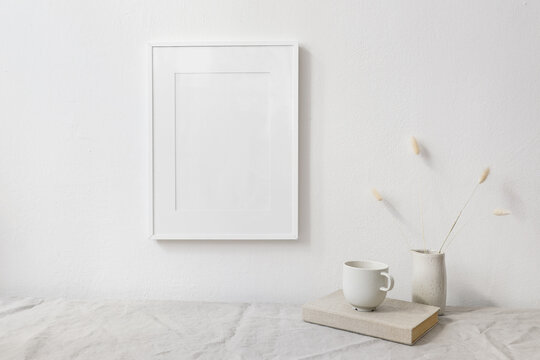 Neutral breakfast still life scene. White wooden picture frame mockup. Vase with dry lagurus bunny tail grass. Cup of coffee on book. Linen table cloth, white wall. Scandinavian interior. Boho style.