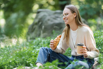 happy woman in glasses holding paper cup and croissant while sitting on grass during picnic.