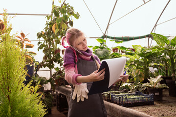 Young woman with pink hair holding tablet for writing and using smart phone for calling to clients in greenhouse of garden mall