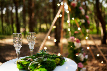 Wine glasses, wedding rings bedding of grass and moss on white table next to bohemian tipi arch...