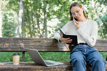 Fototapeta premium cheerful woman in glasses talking on smartphone while sitting near laptop on bench.