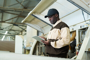 Naklejka na ściany i meble African manual worker in uniform using digital tablet to control machine at factory