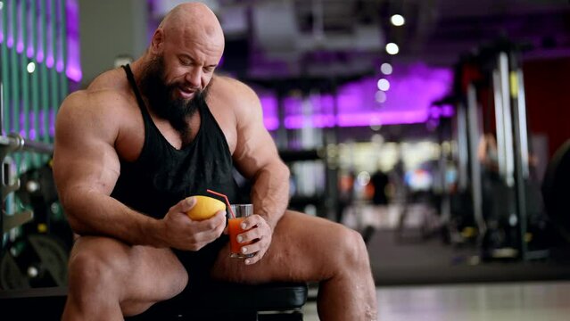 A male bodybuilder with fruit juice relaxes, enjoying after a workout, sitting on the simulator in the gym. An athlete drinks a cocktail