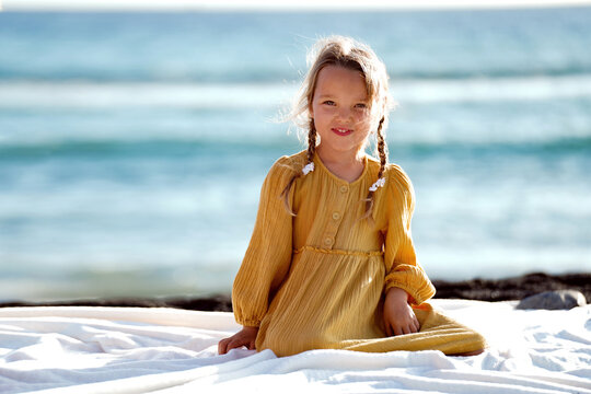 Happy little girl in yellow cute dress enjoying sunny day at the beach, sitting on the blanket and smiling to the camera.