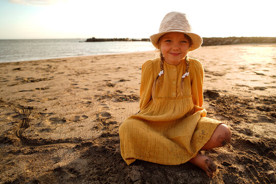 Happy little girl in yellow cute dress and summer hat enjoying sunny day at the beach, sitting on the sand and smiling to the camera.