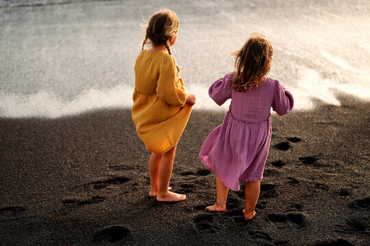 Happy little cute girls enjoying sunny day at the beach, standing on the coast and looking at the waves. Family summer vacation. Sister's love.