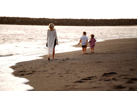 Joyful mother with three kids walking on the beach during sunset. Summer vacations with children. Back view.