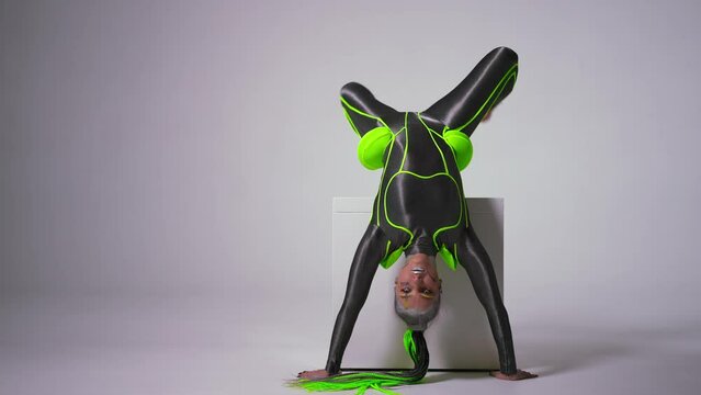 Wide shot flexible gymnast with bright makeup upside down performing on white cube at white background. Portrait of confident fit Caucasian young woman looking at camera dancing