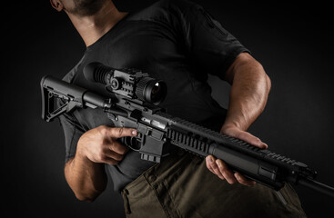 A man holds an automatic carbine with an optical sight and a night vision device in his hands. A fighter in dark clothing with a weapon in his hands. Dark back.